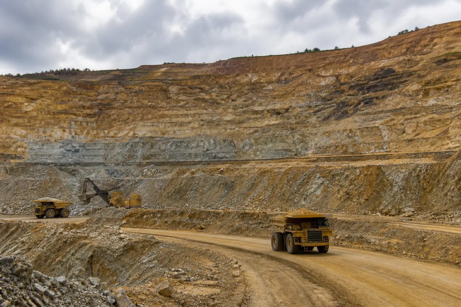 A wide daylight establishing shot of a terraced open-pit gold mine with large haul trucks and an excavator working the benches under an overcast sky.