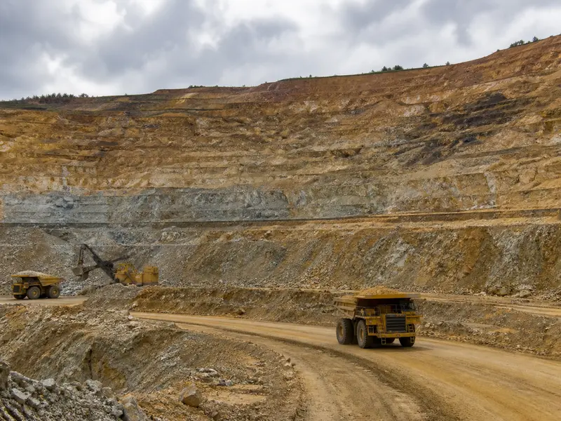 A wide daylight establishing shot of a terraced open-pit gold mine with large haul trucks and an excavator working the benches under an overcast sky.