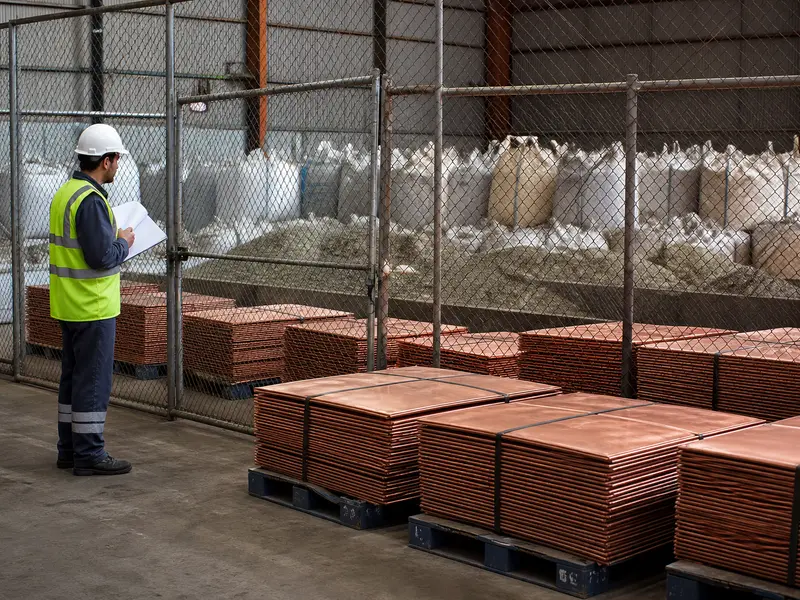 A daylight photograph inside a busy industrial metals warehouse shows neat rows of unlabeled copper cathodes and bulk sacks of mineral concentrates stacked behi