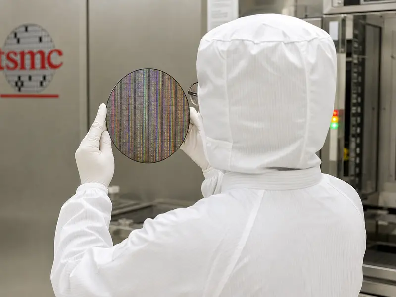 A technician in a full white cleanroom bunny suit inspects a shiny silicon wafer under fab lighting inside a TSMC semiconductor plant, the company's logo faintl
