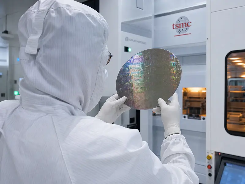 A technician in a full white cleanroom bunny suit inspects a polished silicon wafer inside a TSMC semiconductor fab, framed over the shoulder under cool cleanro