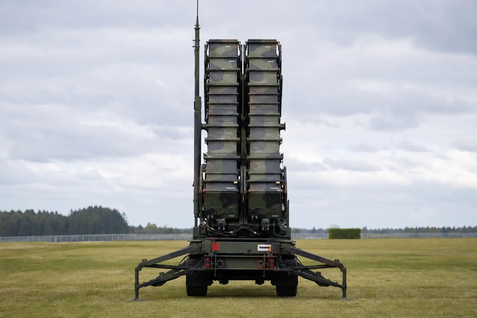 A Patriot missile defense launcher stands in an open field under flat daylight, photographed straight-on with its launch tubes raised toward the sky.