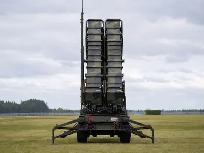 A Patriot missile defense launcher stands in an open field under flat daylight, photographed straight-on with its launch tubes raised toward the sky.