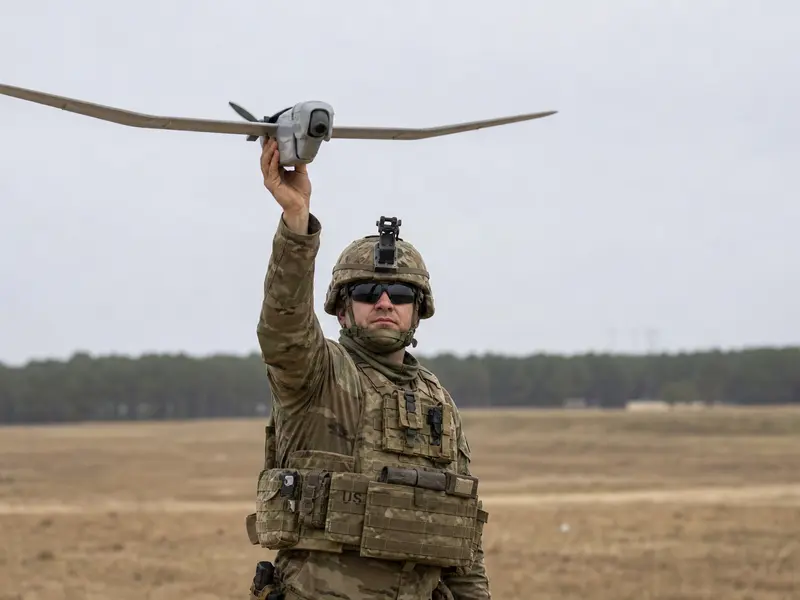 U.S. Army soldiers in fatigues launching a small hand-held reconnaissance drone during a training exercise on an open field under flat daylight.