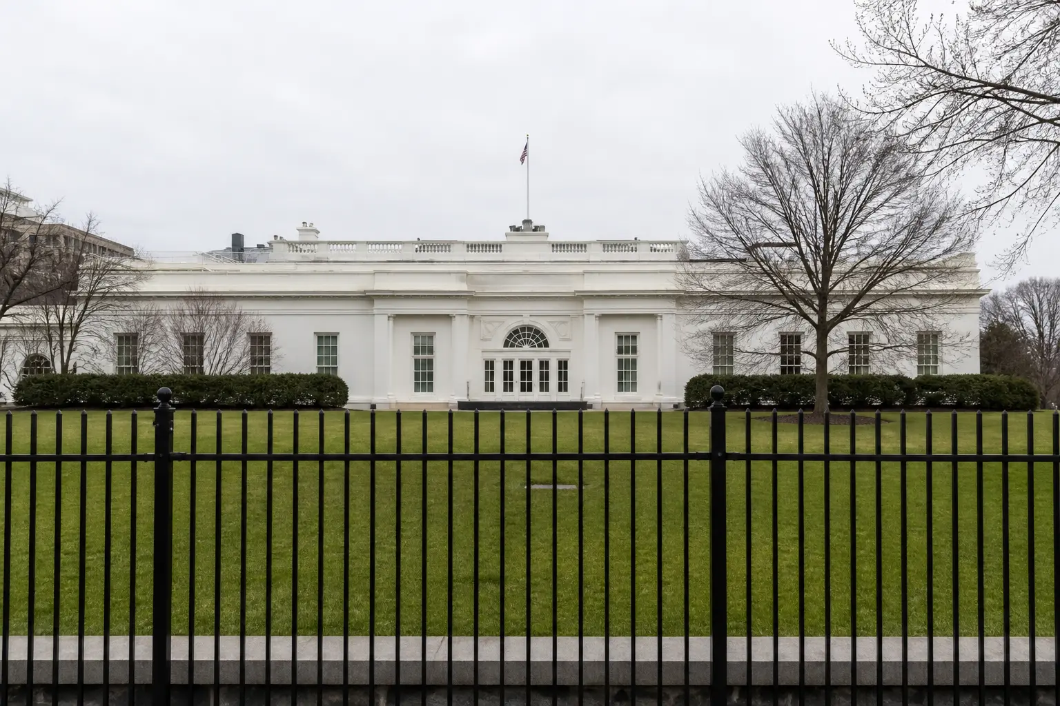 A wide front-on shot of the West Wing of the White House in flat daylight, seen across the lawn behind the perimeter fence.