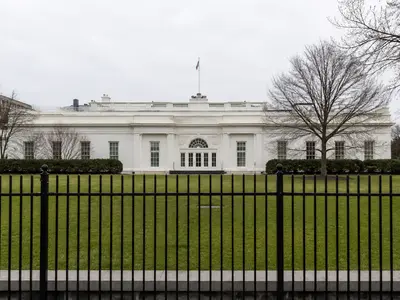 A wide front-on shot of the West Wing of the White House in flat daylight, seen across the lawn behind the perimeter fence.