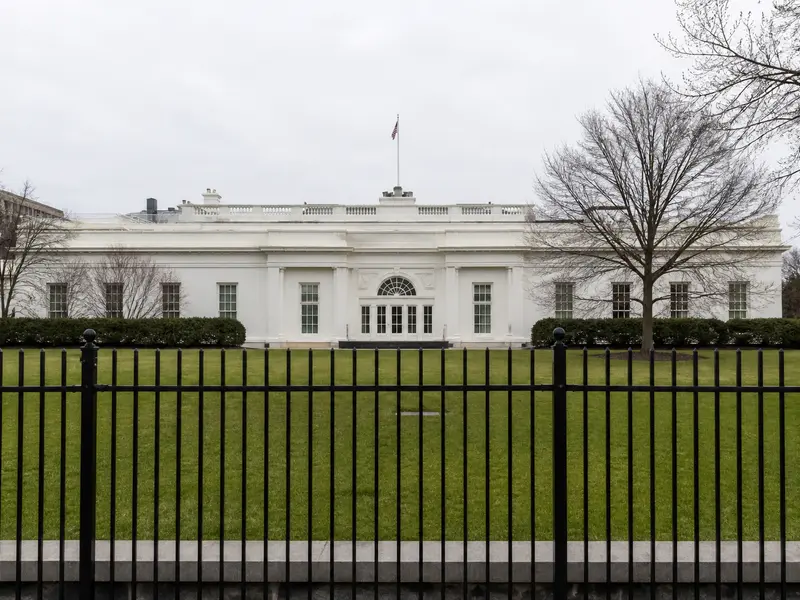 A wide front-on shot of the West Wing of the White House in flat daylight, seen across the lawn behind the perimeter fence.
